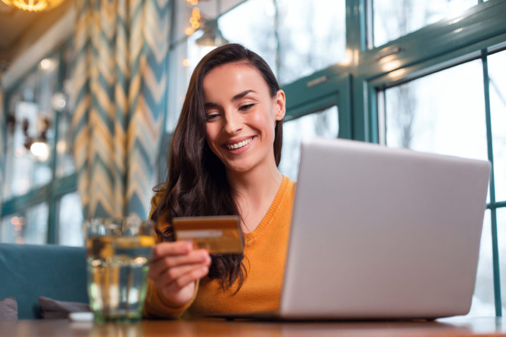 Irresistible positive woman using credit card Advanced payment. Optimistic energetic calm woman using credit card while gazing down and laptop standing on table